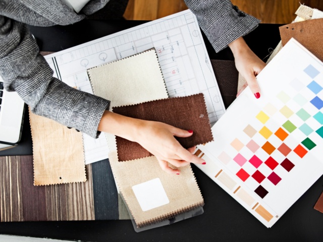 a person holding a piece of paper with colours over a table. Fabric samples can also be seen in the table. 