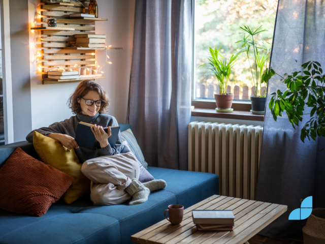 A woman sat in the sofa reading a book, the picture also contains a shelf with books and fairy lights and a coffee table.