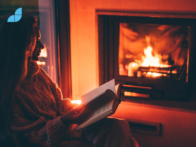A woman sat in front of a fireplace reading a book.