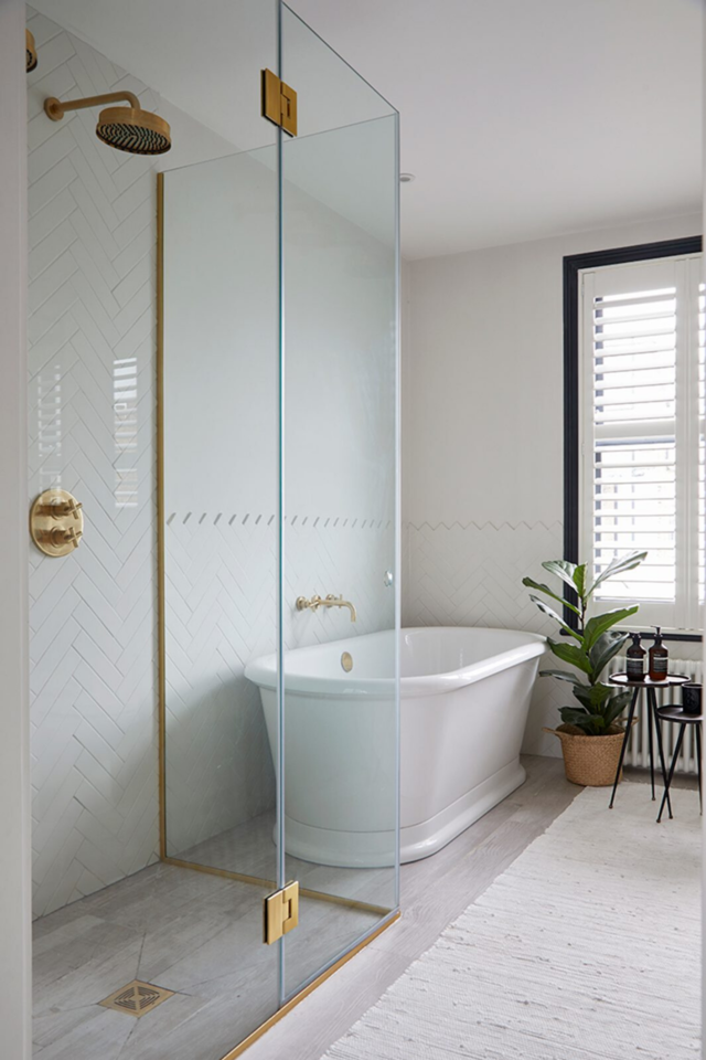 A bathroom with a white bath and gold shower. White walls and natural light coming inside of the room. A plant can also be seen on one of the sides. 